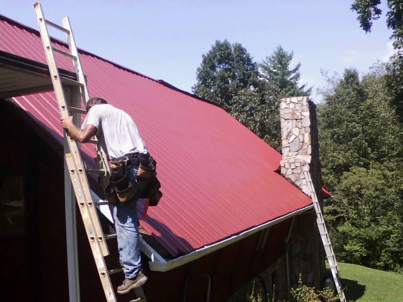 Roofer on a ladder beside a red metal roof and stone chimney — Soffit & Fascia Repair by Roofing Brothers in Ranchita